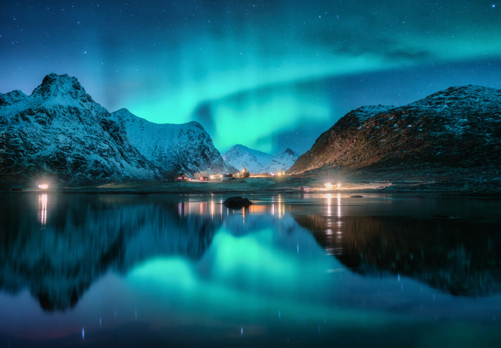Aurora borealis, snowy mountains, sea, fjord, reflection in water, street lights at starry winter night. Lofoten, Norway. Northern lights. Landscape with polar lights, snowy rocks, sky with stars